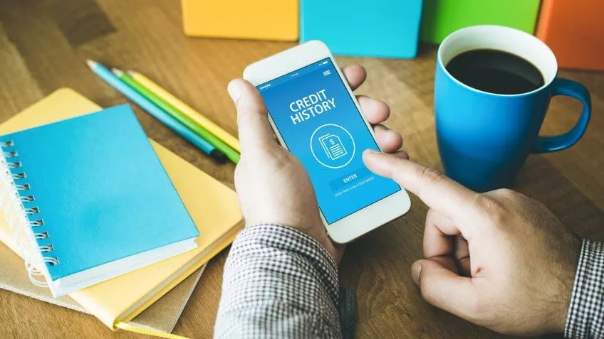 man checking his credit history on his smart phone with desk work utensils around him