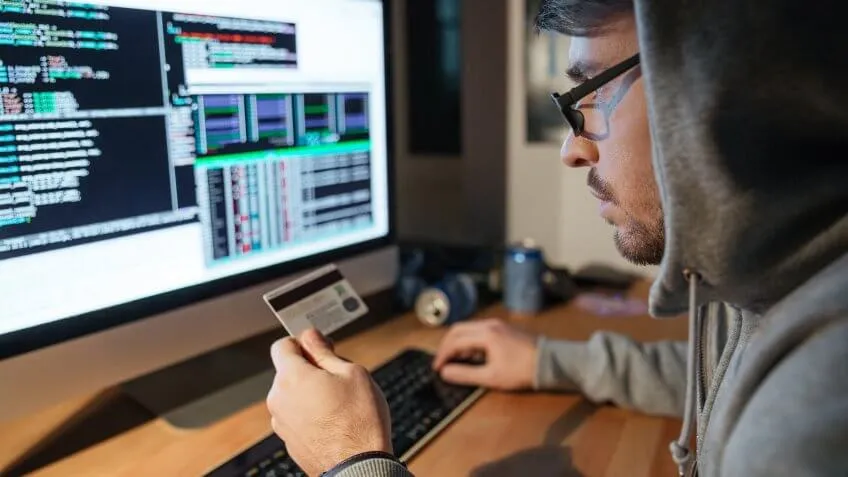Concentrated young hacker in glasses stealing money from diferent credit cards sitting in dark room.