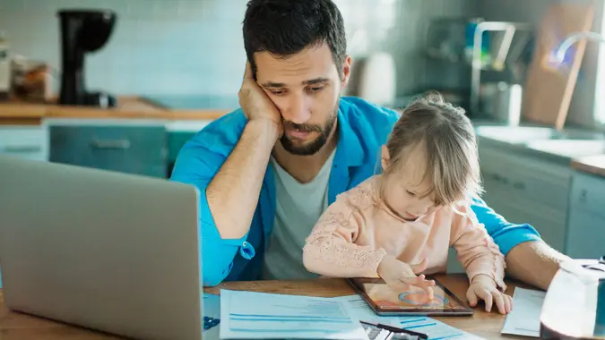 Photo of a father and daughter sitting in kitchen,father is doing home financials while daughter is having fun with digital tablet.