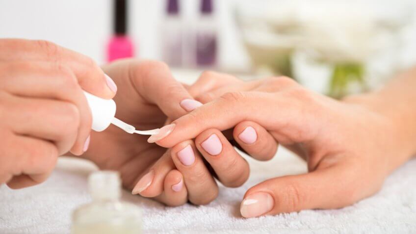 manicurist doing manicure client painting nails with transparent nail polish in salon on white towel.