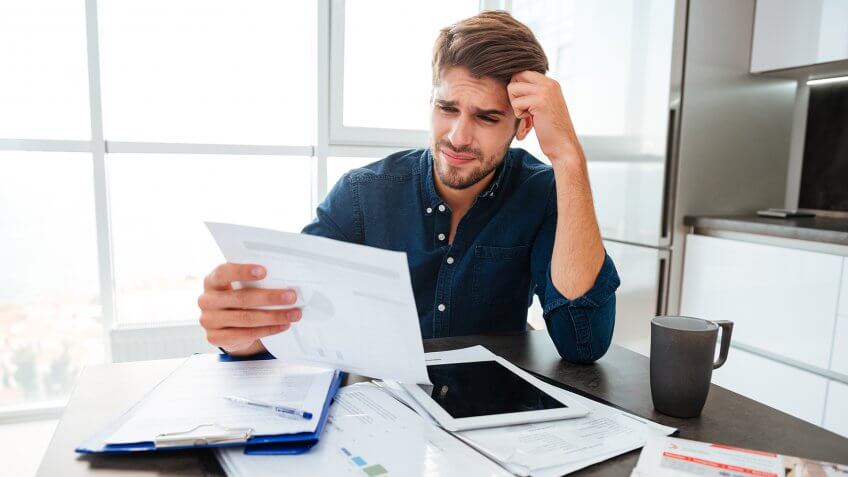 Young confused man analyzing finances at home while holding head with hand and looking at documents.