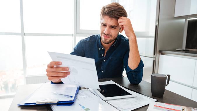 Young confused man analyzing finances at home while holding head with hand and looking at documents.