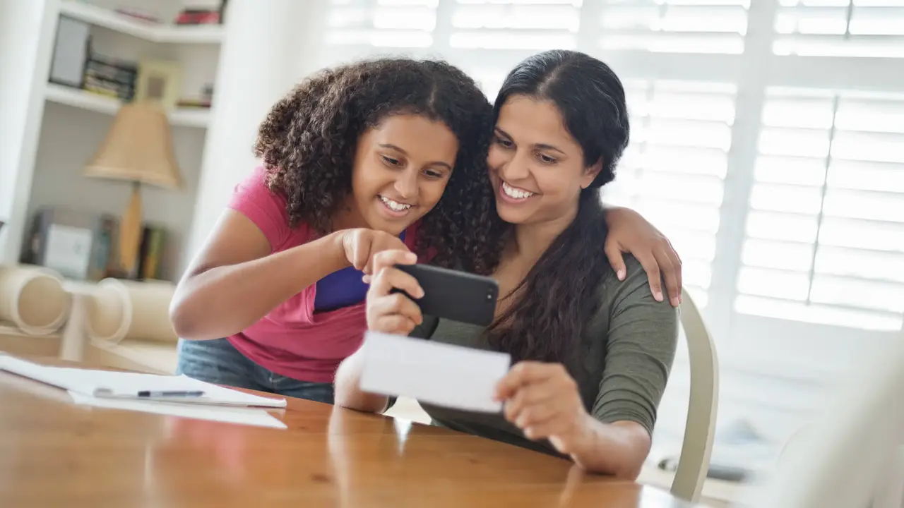 Happy teenage daughter and mother depositing check through smart phone at home.