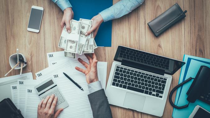 usiness woman giving packs of dollars to her accountant and paying taxes, he is examining tax forms and calculating expenses.