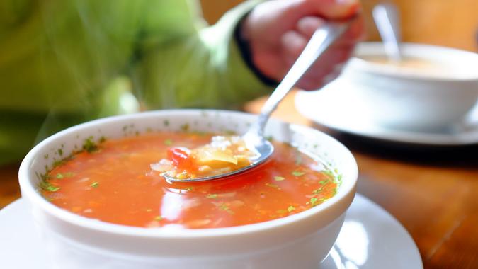 Bowl of hot vegetable soup and hand holding spoon.