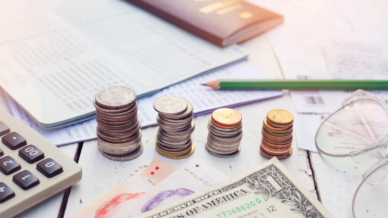 Picture of coin stacks placed on a desk, surrounded by multiple notebooks, paper bills, pencils and a calculator