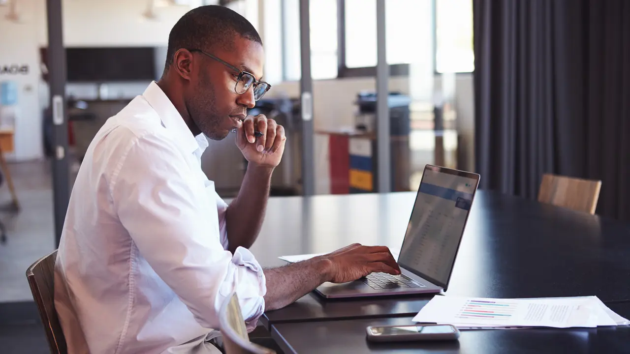 A trader evaluates his portfolio with a laptop and paperwork, analyzing stock charts and financial data.