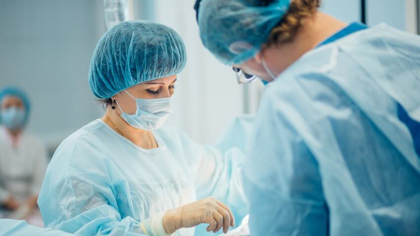 22 Industries That Pay Less (and More) Than They Did 10 Years Ag, two surgeons at work in operating room. close up
