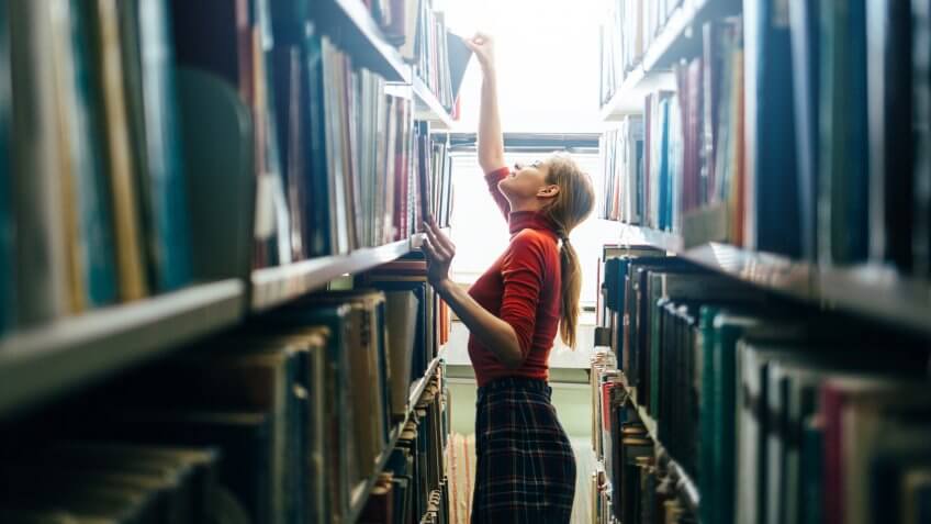 22 Industries That Pay Less (and More) Than They Did 10 Years Ag, Woman taking book from library bookshelf. Young librarian search