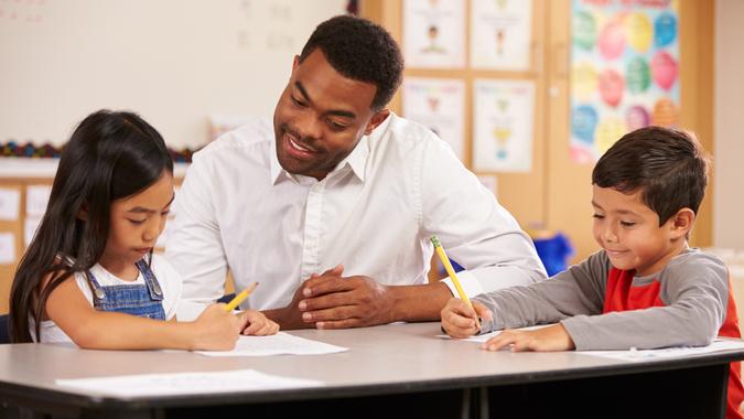 96 Money-Making Skills You Can Learn in Less Than a Year, Teacher sitting at desk with two elementary school pupils