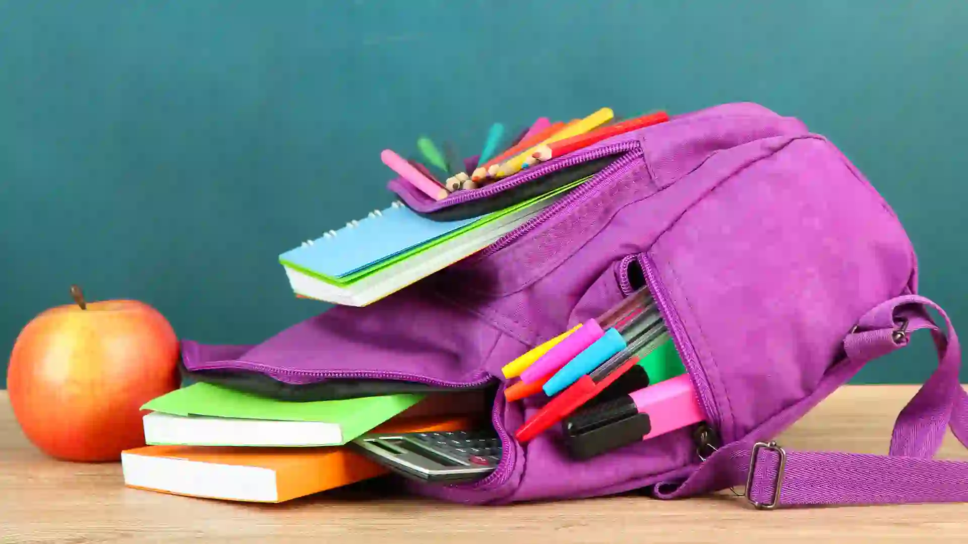 Purple backpack with school supplies on wooden table on green desk backgroundPurple backpack with school supplies on wooden table on green desk backgroundPurple backpack with school supplies on green desk background.