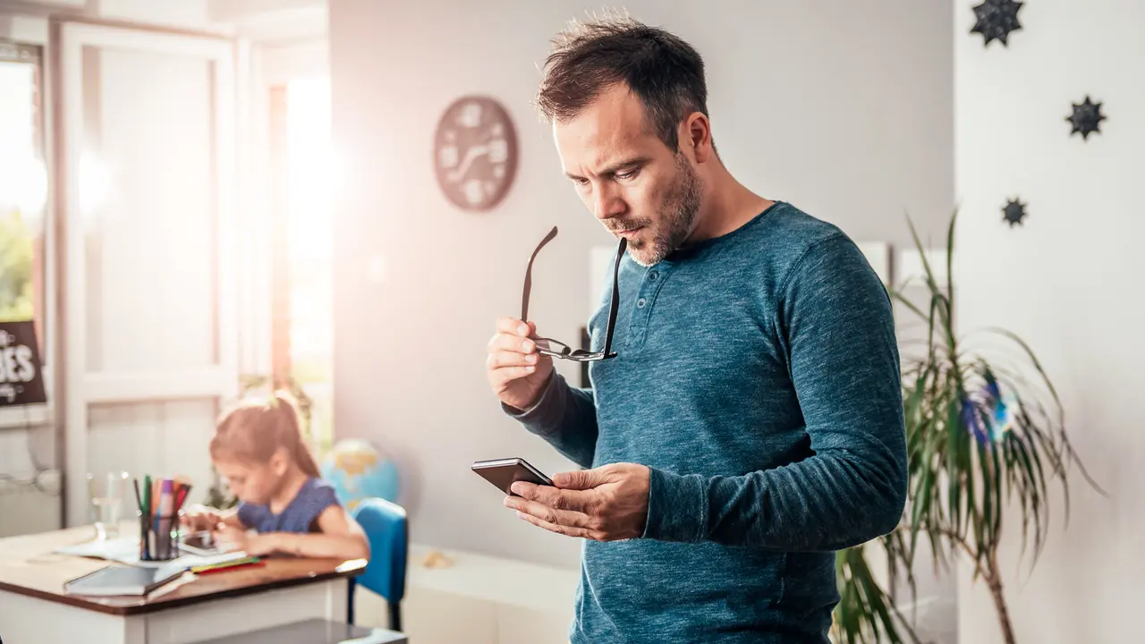 Worried father looking at smart phone with eyeglasses in his hand, in background his daughter doing homework.