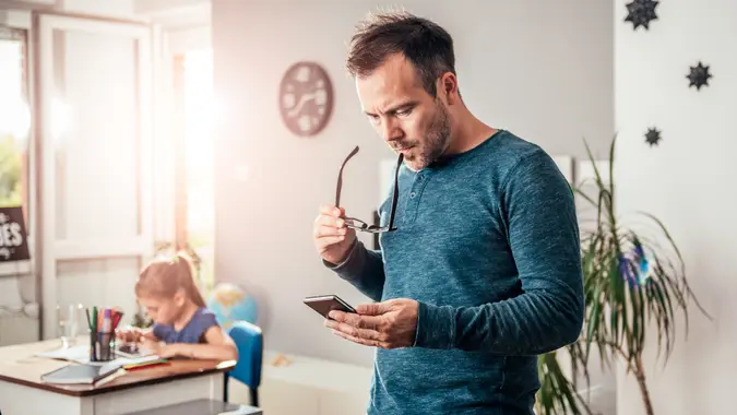 Worried father looking at smart phone with eyeglasses in his hand, in background his daughter doing homework.