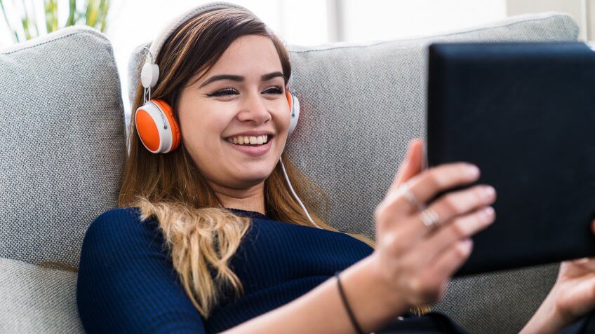 Young adult woman wearing headphones watching TV on a digital tablet whilst relaxing on the sofa.