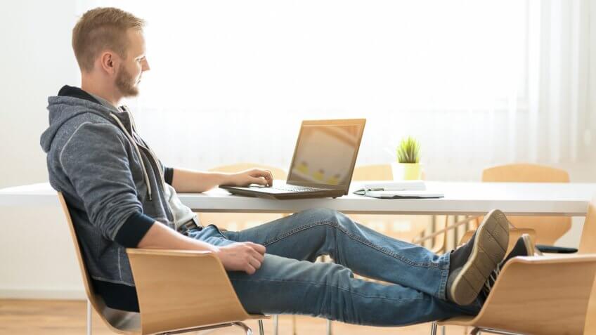 Young man working casual at his laptop with legs on the chair.