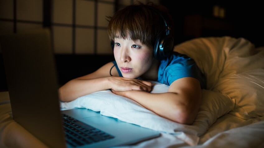 Mid adult woman wearing headphones lying in bed watching tv on her laptop.