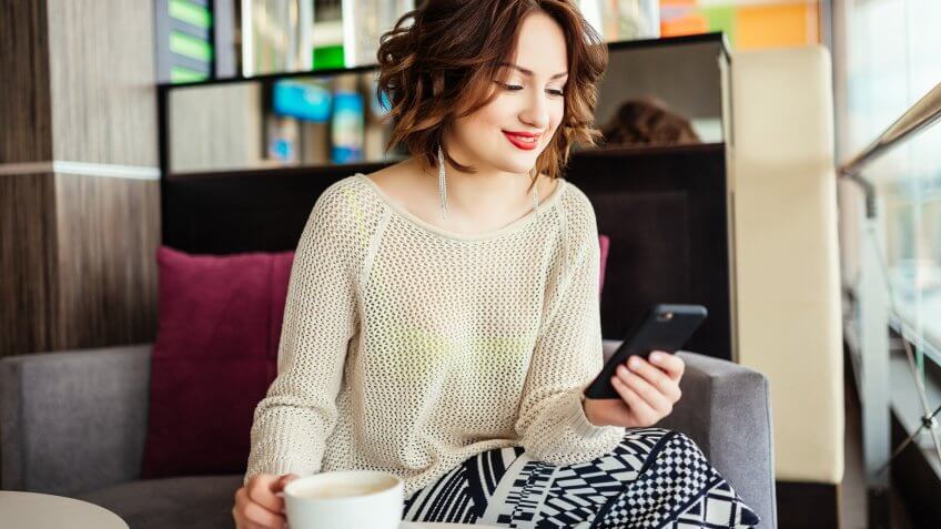 Smiling young business woman sitting in cafe at table and use smartphone.