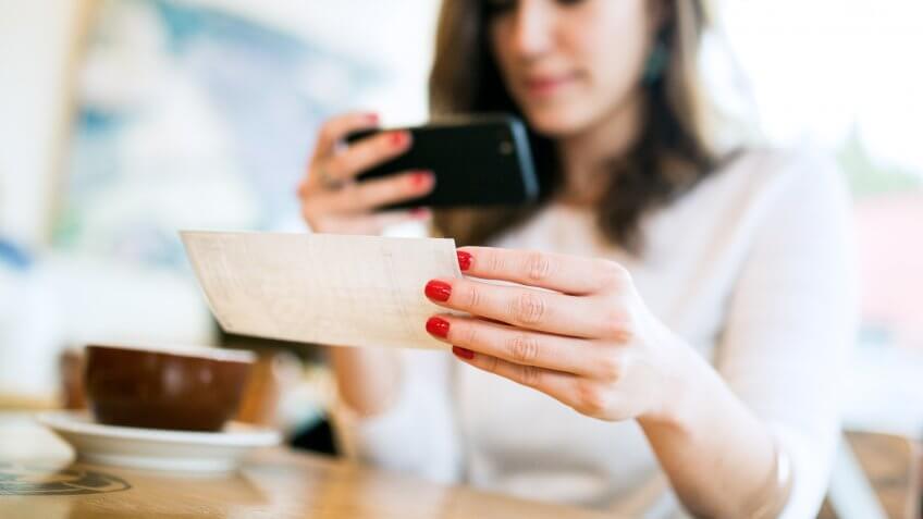 11730, 20-29 Years, A smiling young woman takes a picture with her smart phone of a , Bank Deposit Slip, Cafe, Check - Financial Item, Coffee Shop, Currency, E-commerce, Electronic Banking, Horizontal, Paycheck, Paying, Photographing, Smart Phone, Smiling, Women, Young Adult, also known as "Remote Deposit Capture". She sits in a coffee sh, depositing, enjoying an espresso latte. Bright sunlight shines in the windo, people