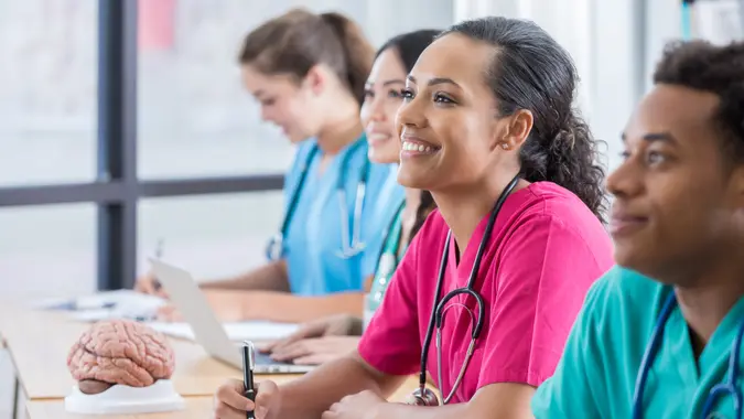 Im Mittelpunkt steht eine Krankenpflegestudentin, die inmitten ihrer drei Klassenkameraden lächelt, während zwei ihrer Klassenkameraden zum Professor aufblicken.