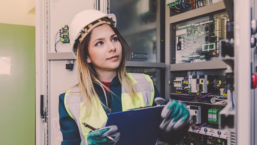 Young woman industrial service engineer wearing white worker headwear standing, working, checking conducts a safety check front of a control panel read and writing notes about technical problems in boiler energy control room of modern thermal or nuclear power plant electric energy station.