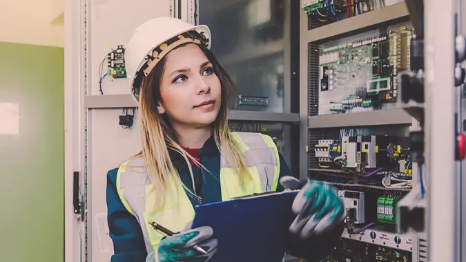 Young woman industrial service engineer wearing white worker headwear standing, working, checking conducts a safety check front of a control panel read and writing notes about technical problems in boiler energy control room of modern thermal or nuclear power plant electric energy station.