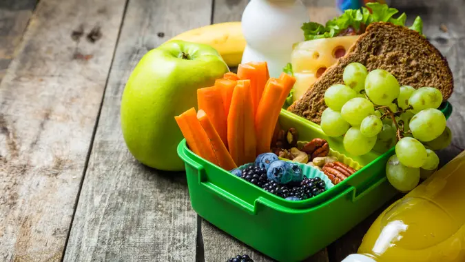 A green lunch container with carrots, grapes, berries and a sandwich on a wood table, with an apple and drink next to it.