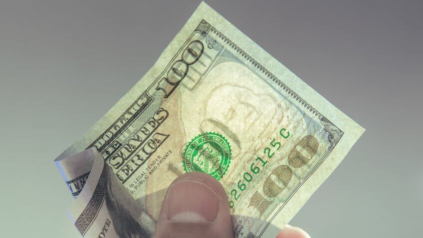 A hand holding a one hundred dollar bill with translucent Franklin watermark visible.