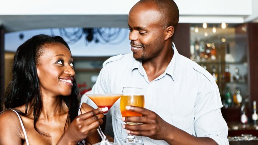 A romantic-looking young couple smile and toast each other in cocktails at an upmarket bar.