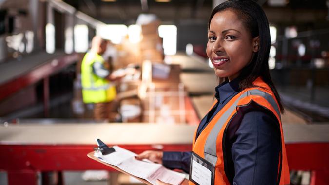 Portrait of a warehouse manager holding a clipboard with workers in the background.