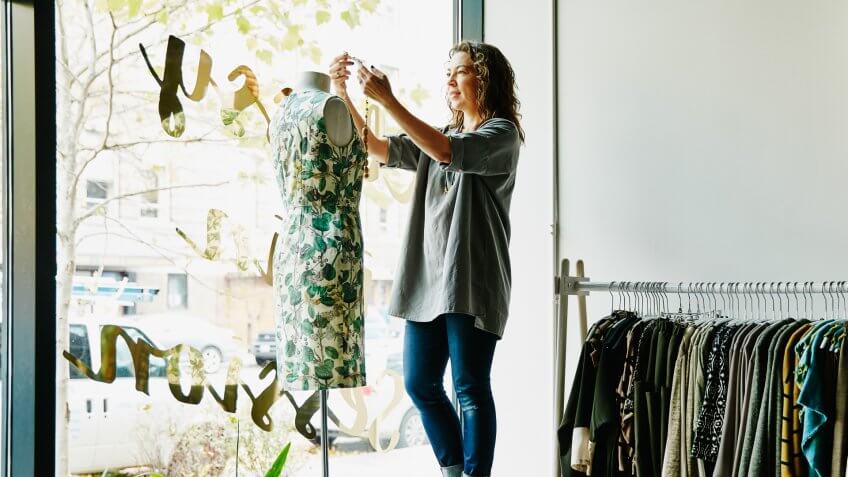 Female business owner dressing dress form in window of boutique.