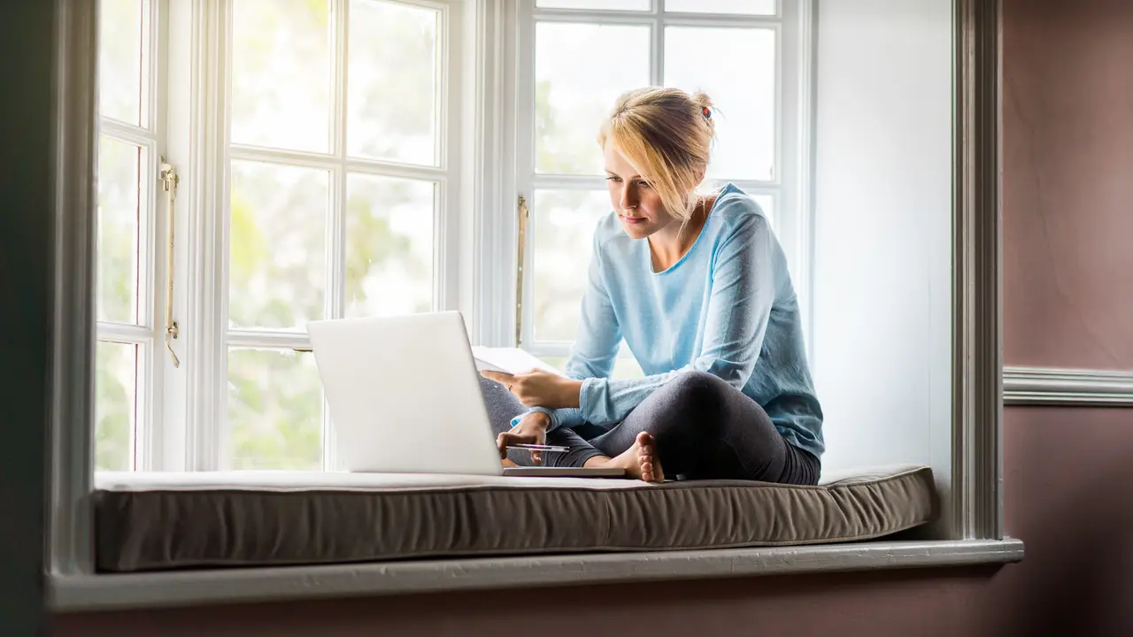 A full length photo of woman using laptop while holding documents at home.