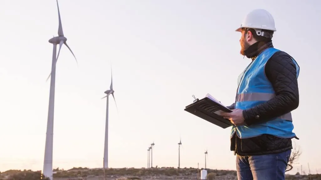 Young Handsome Beard Male Engineer in Wind Mill Power Generator Station with white safety hat and wind turbines on background.