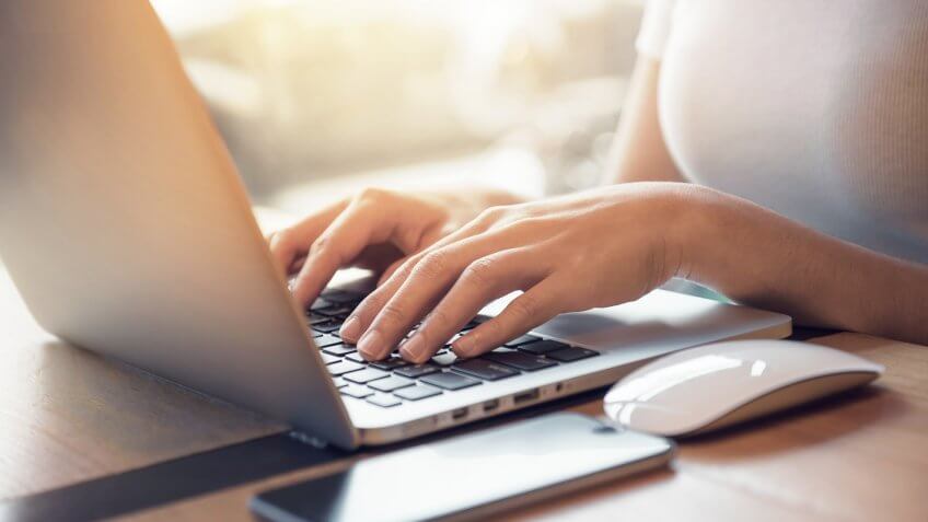 Closeup woman's hands typing on a laptop that is on a wooden desk with a mobile smartphone and mouse computer.