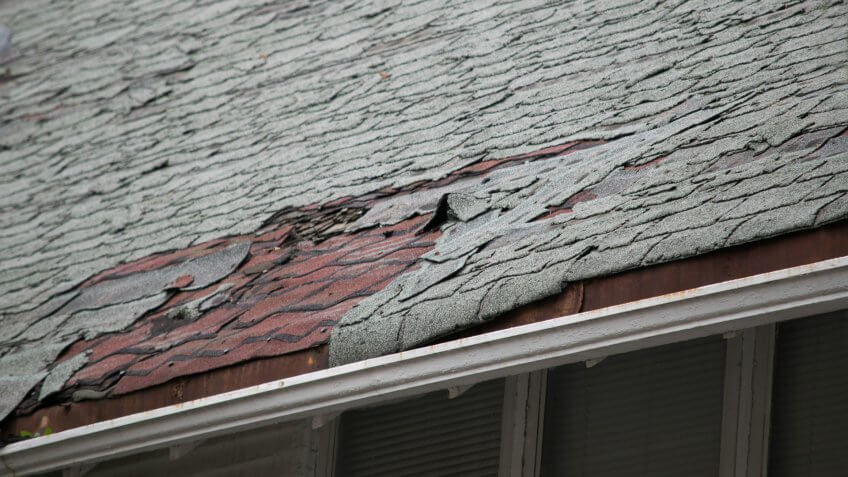 Roof of a residential house showing damage to the tile and gutter systems, multiple layers of shingles, missing shingles, and rusty gutters.
