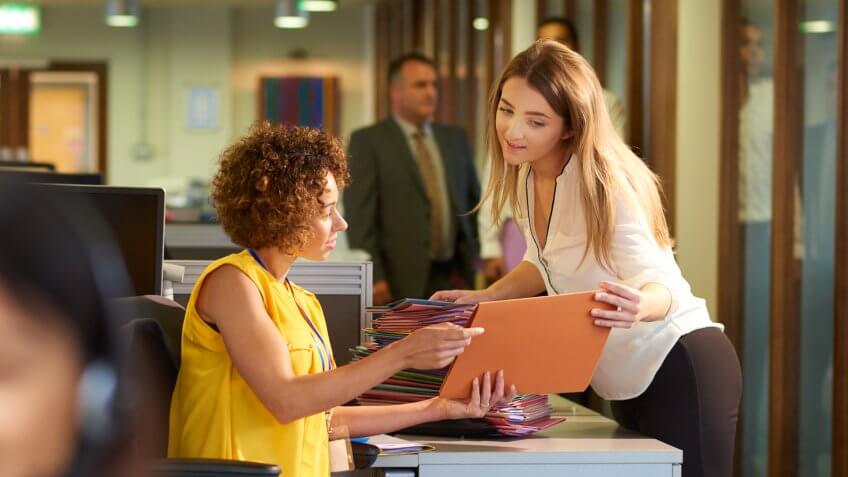 a young office junior takes a file from her supervisor for filing in a large open plan office .