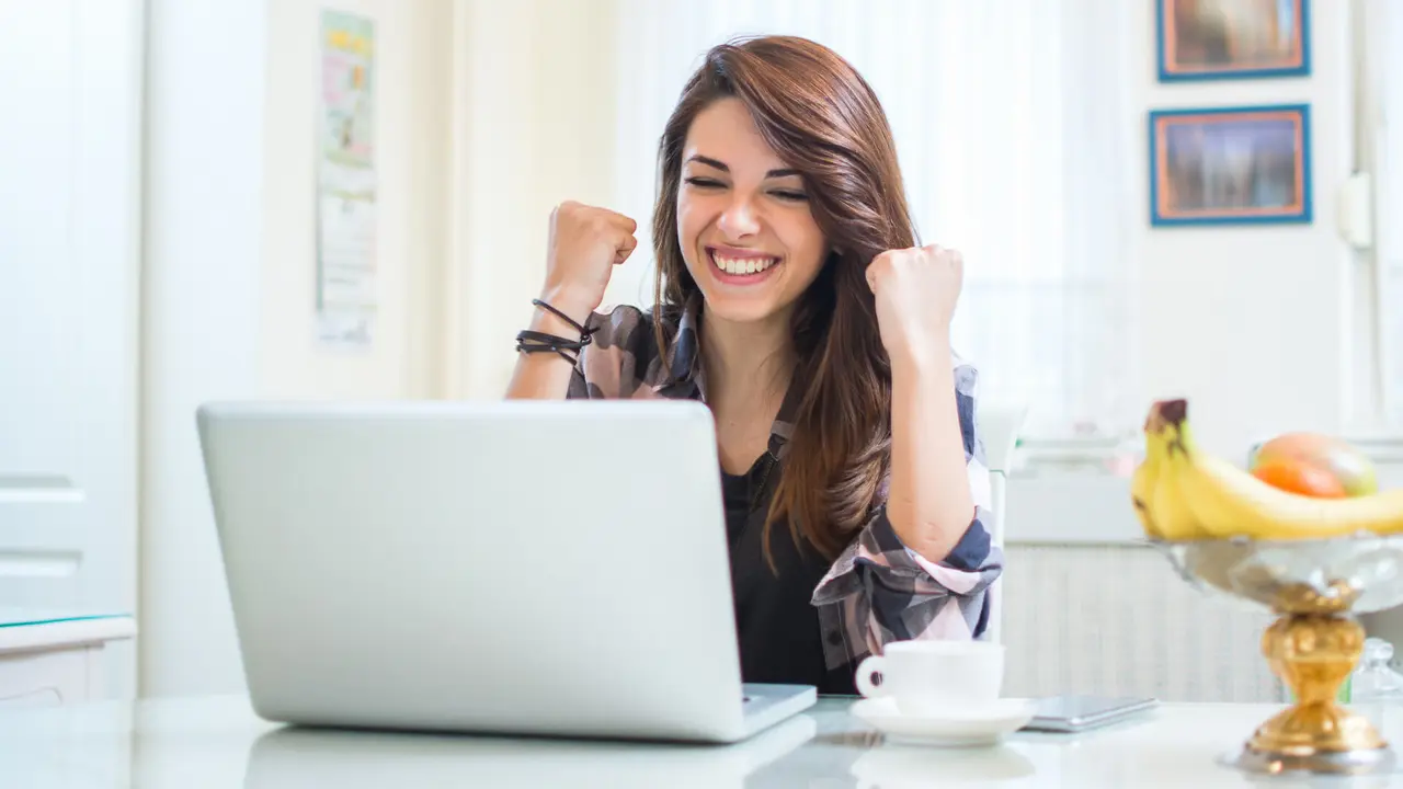 Portrait of happy young woman celebrating success with arms up in front of laptop at home.