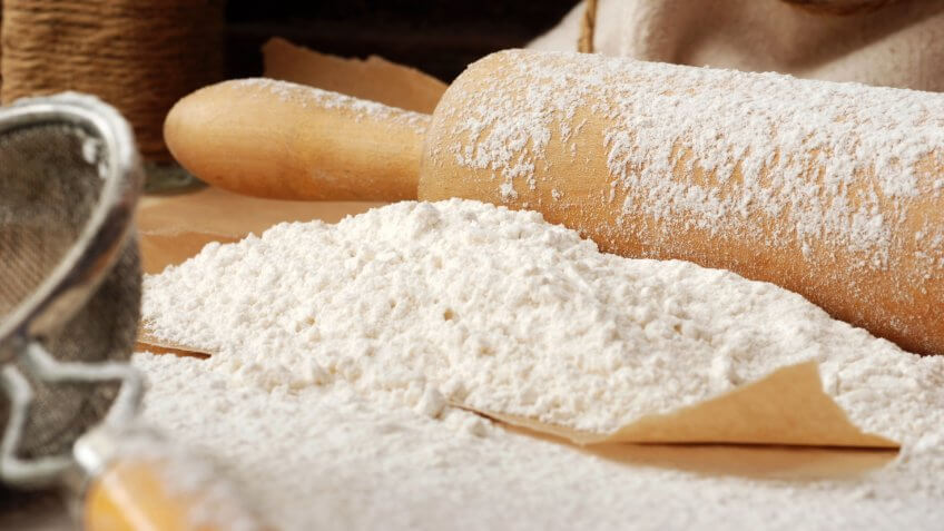 Baking still-life with flour heaped onto parchment paper, vintage wooden rolling pin, vintage sieve, and canvas flour sack with rope in background.