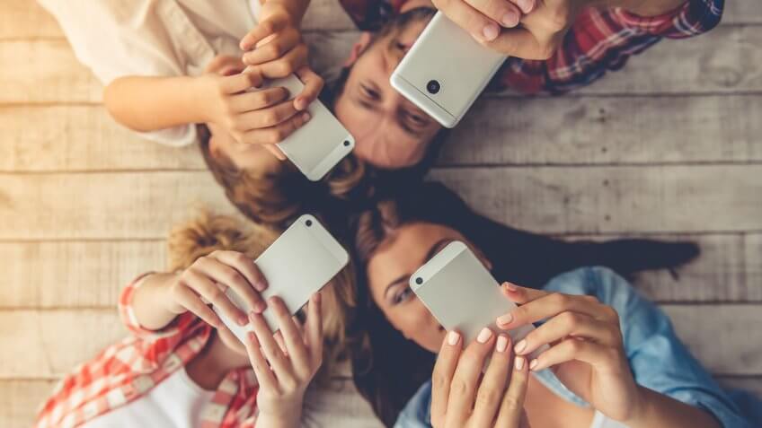Top view of young parents and their children using smartphones and smiling while lying together on the floor at home.