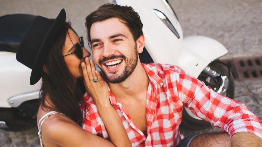 Close up of attractive young couple whipsering to ear a secret while sitting near scooter and smiling.
