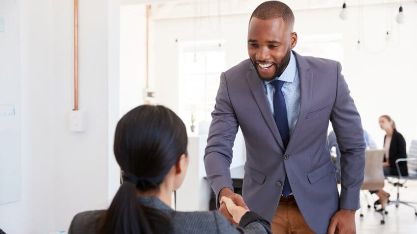 Black businessman and seated woman shaking hands in office.