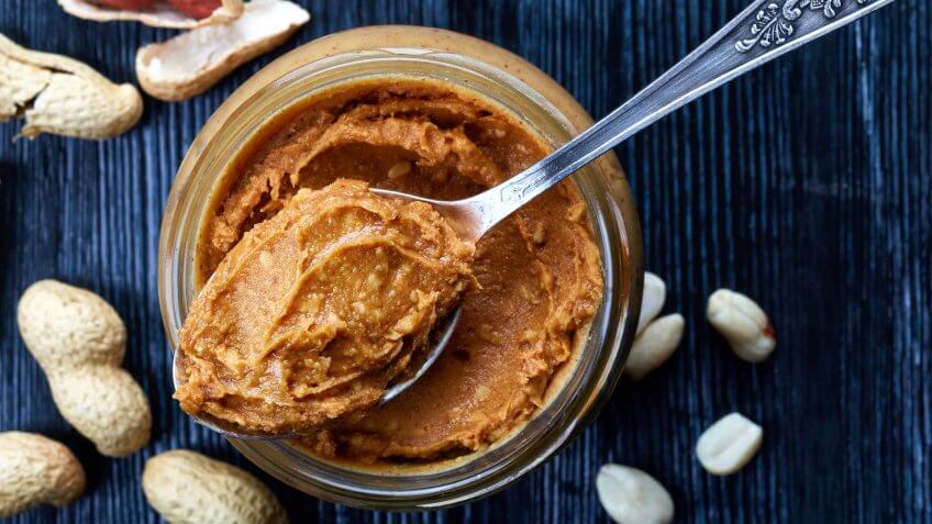 Jar and spoon of peanut butter and peanuts on dark wooden background from top view.