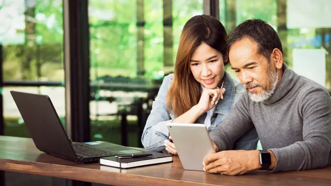 Smiling mature man with white stylish short beard using tablet with charming woman teenage.