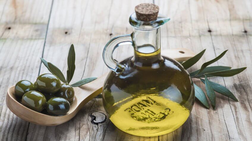 A glass bottle of olive oil and a wooden spoon with olives on a table.