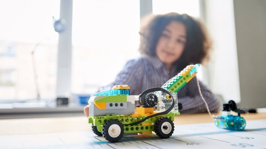 Close-up shot of toy lifting truck and smaller car made of plastic building blocks following blue line one after another, teenage boy looking at them with smile in classroom.