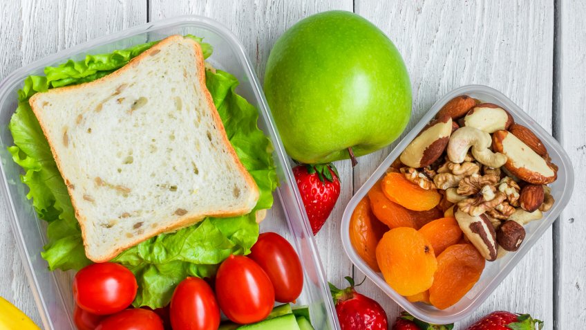 school lunch boxes with sandwich and fresh vegetables, bottle of water, nuts and fruits on white wooden background.