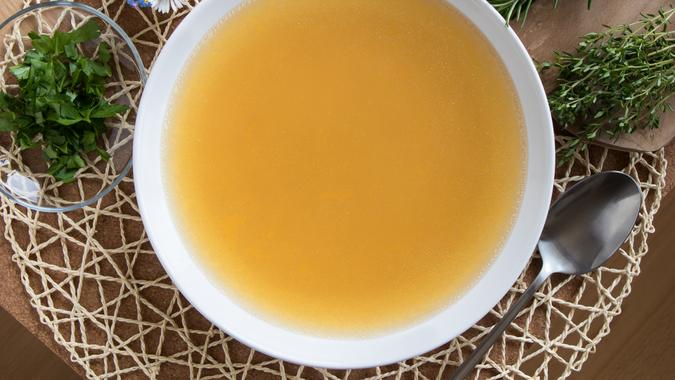 Bone broth made from chicken, served in a white plate, with chopped parsley, green herbs, and flowers in the background.