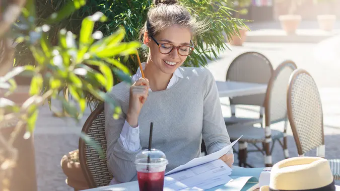 Outdoor portrait of young good-looking Caucasian female in glasses sitting at cafe table outdoors and filling in forms on papers with pencils feeling happy about both working and having nice rest.
