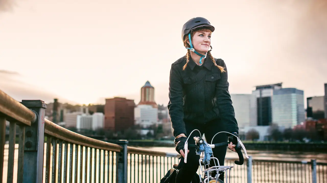 A smiling young woman commuting in an urban city environment on her street bicycle, waterproof panniers on her bike rack.
