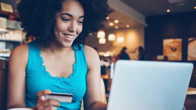 Smiling young African ethnicity woman sitting in cafe, holding a credit card and typing on a laptop.