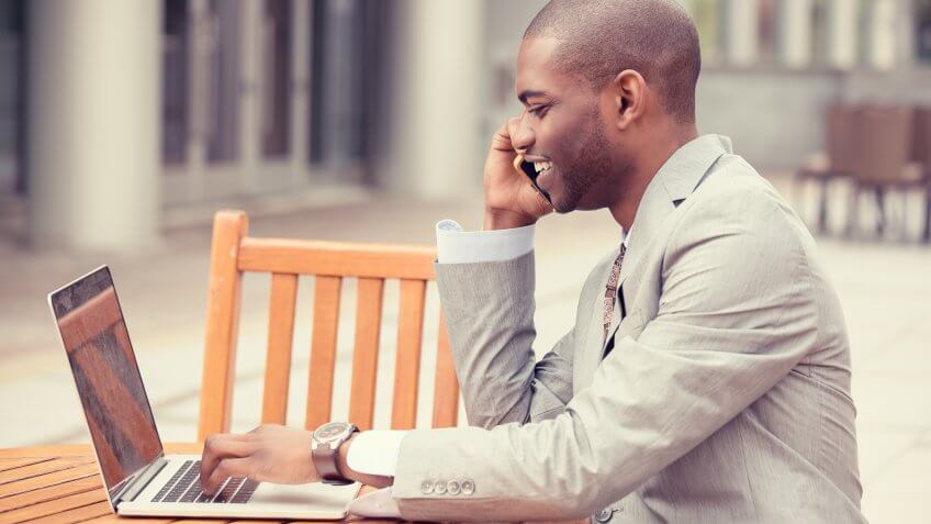 Side profile handsome young businessman working with laptop outdoors talking on mobile phone.